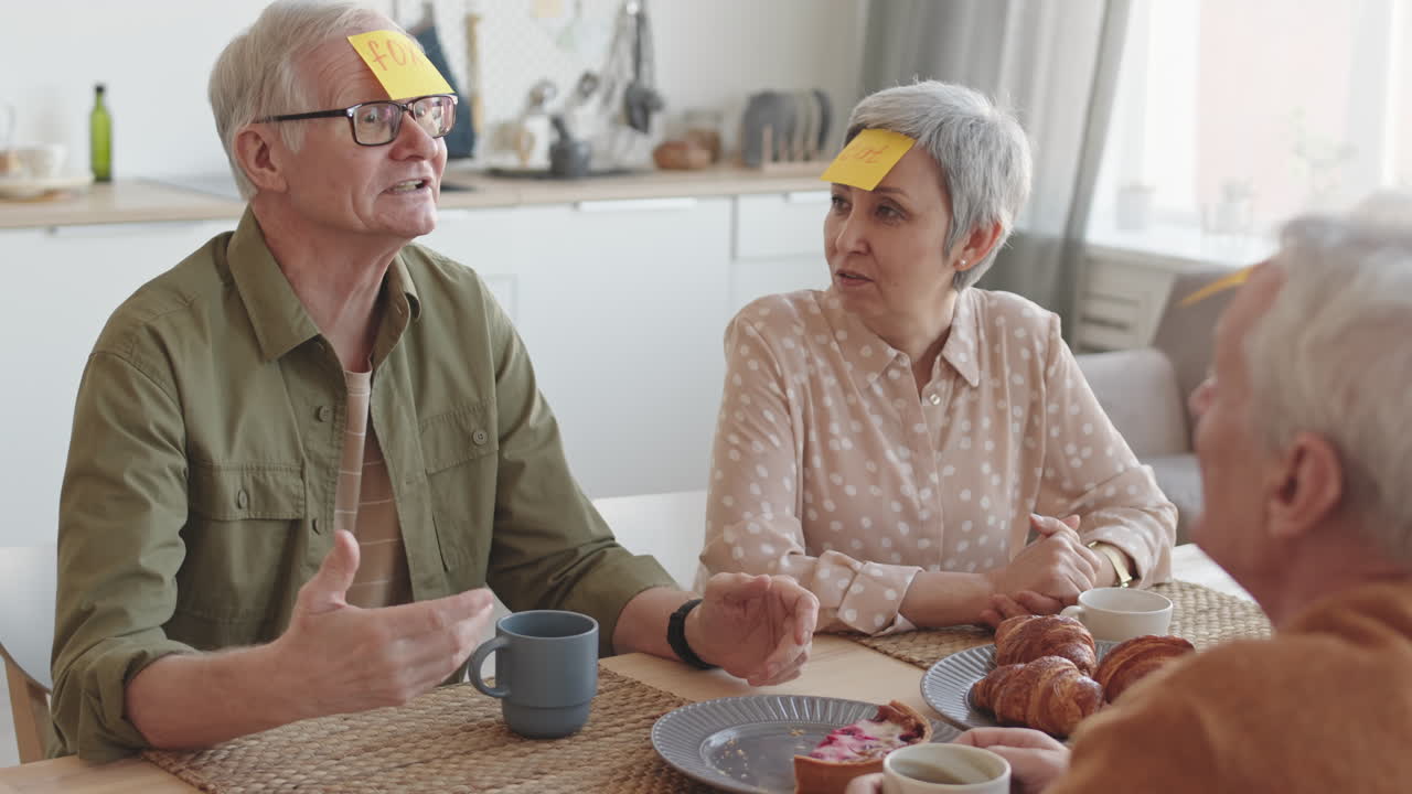 Waist-up of grey-haired people sitting at table, playing Forehead Detective game with yellow sticky notes on head, Asian woman having cat written on it, Caucasian man winning, taking fox note off