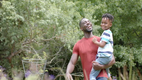 African-American-father-carries-his-son,-both-smiling-in-a-lush-garden-at-home-with-copy-space