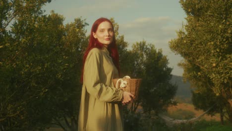 woman with red hair in a green dress holding a basket in an orange grove