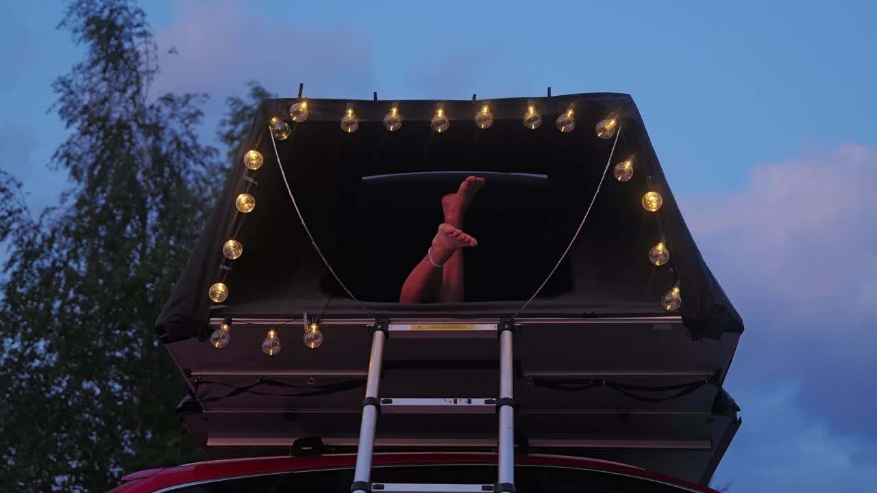 Female legs playfully swing from rooftop tent lit with bulbs during twilight