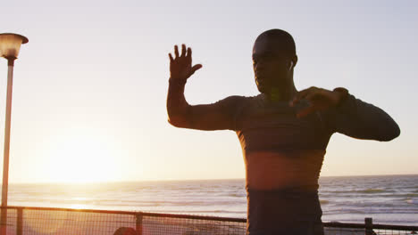 Focused-african-american-man-stretching,-exercising-outdoors-by-seaside-at-sunset