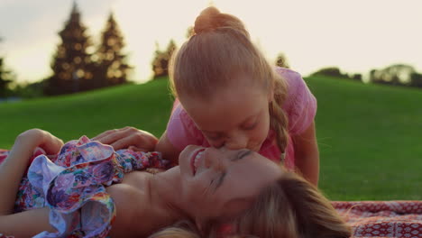 woman laughing with girl at meadow. daughter kissing mother in city park.