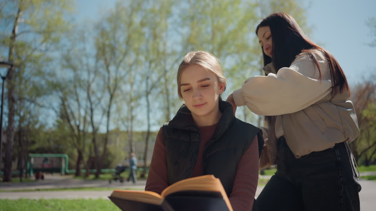 estudiante caucásico asiático leyendo en el parque, ayudando con el pelo y la postura, descanso de estudio tranquilo con un libro en el regazo, luz cálida de primavera y abedules, amistad de apoyo y concentración silenciosa en las páginas, ambiente relajado en el campus