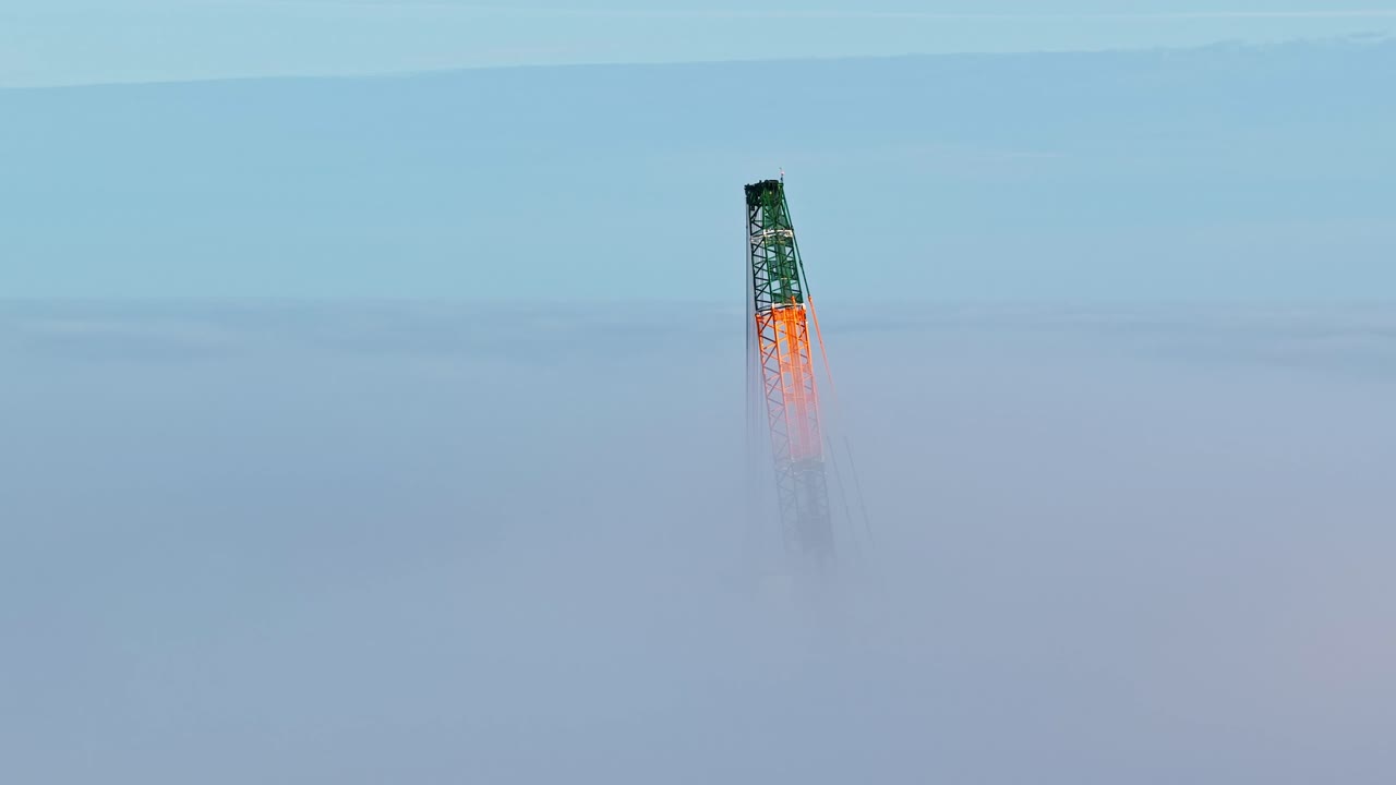 The green and orange top of a tall crane rises above the mist