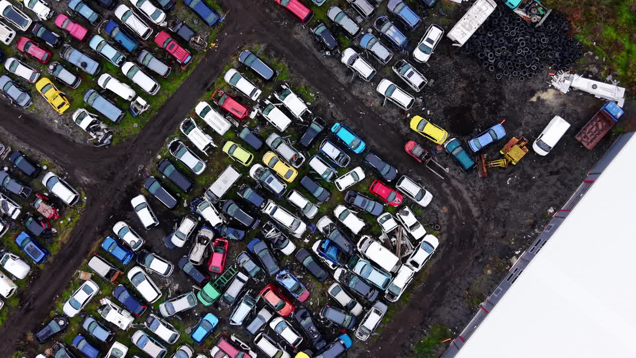 Aerial perspective of a vehicle dismantling yard featuring various car models, tires, and heavy machinery for metal recycling and parts recovery. Car salvage lot and tire storage