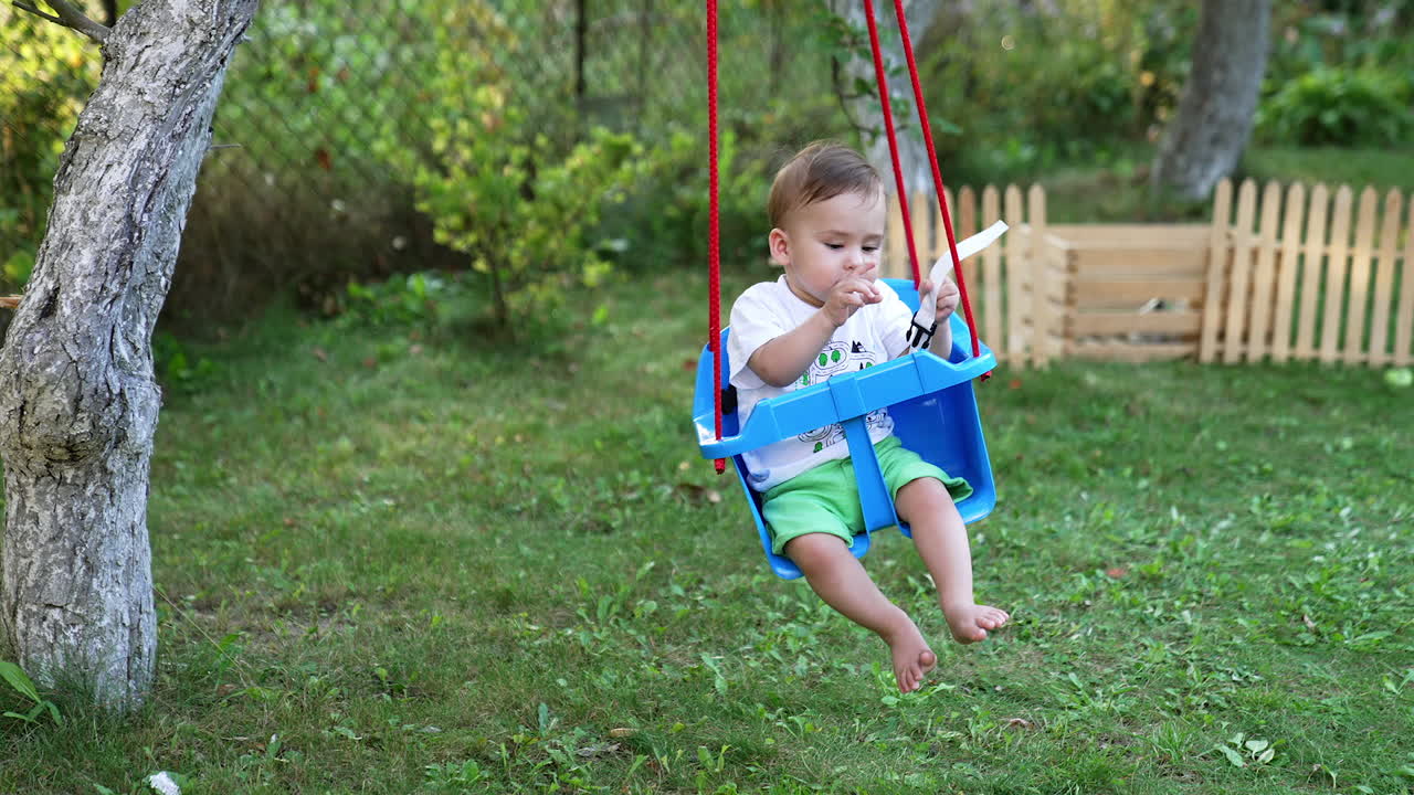 Peaceful Caucasian baby boy swaying in the blue swing in the garden. Trees and wooden fence at backdrop.