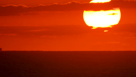 A-cargo-ship-travels-across-in-the-distance-across-the-ocean-horizon-under-a-huge-rising-sun,-glowing-the-sky-bright-orange