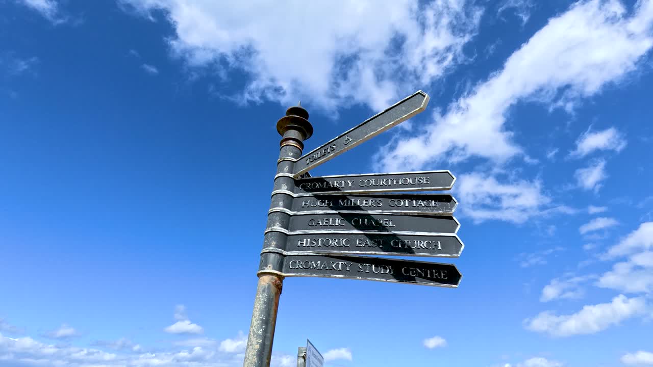 A metal directional signpost slowly rotates under bright daylight, set against a vivid blue sky with scattered clouds in a quiet town environment