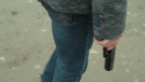 close view of a person in a gray jacket, black canvas and blue jeans holding a handgun by their side, standing on a dirt road, with a blurred background