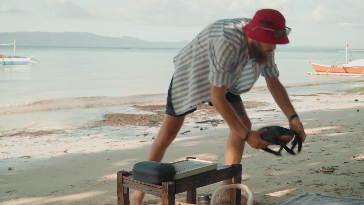 Man preparing tea on beach, setting kettle on low bench, pouring hot water into cup, gentle waves and boats in background, warm morning light, casual striped shirt and red hat, relaxed ritual mood