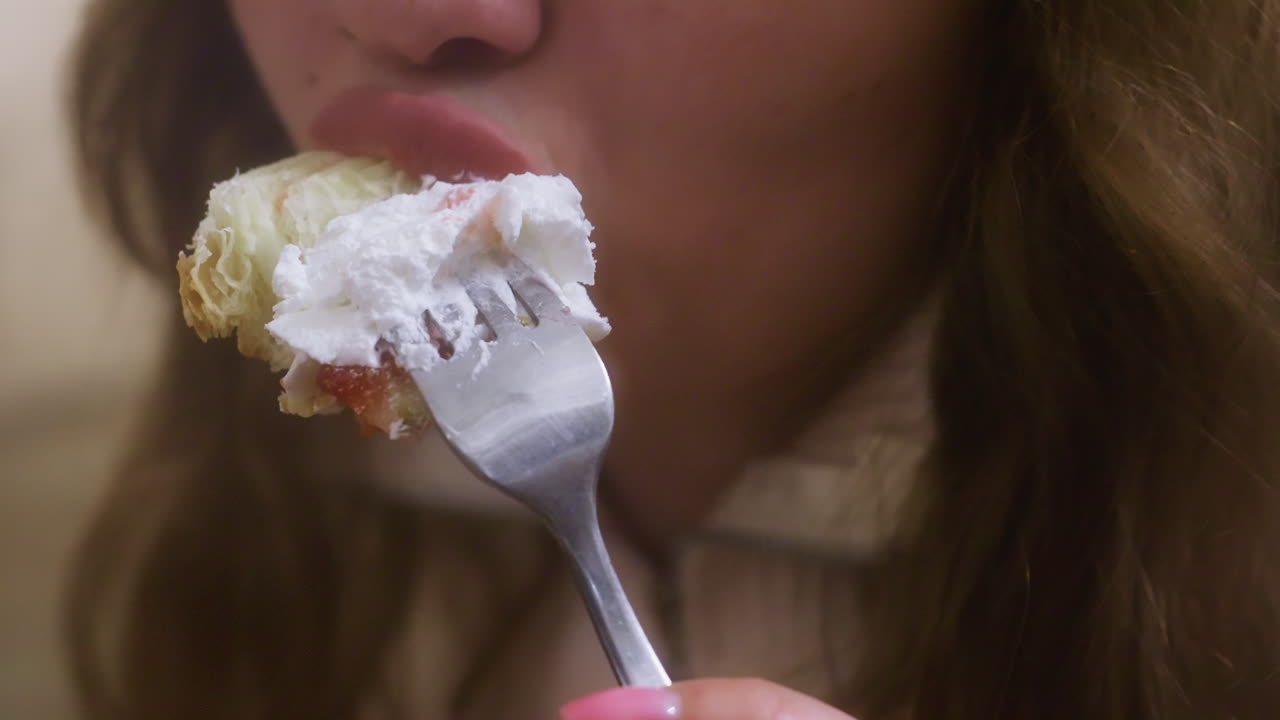 Close-up of hand holding fork and knife, cutting croissant with whipped cream and fruit topping in cozy cafe. The relaxed, enjoyable moment of eating in a soft, inviting atmosphere
