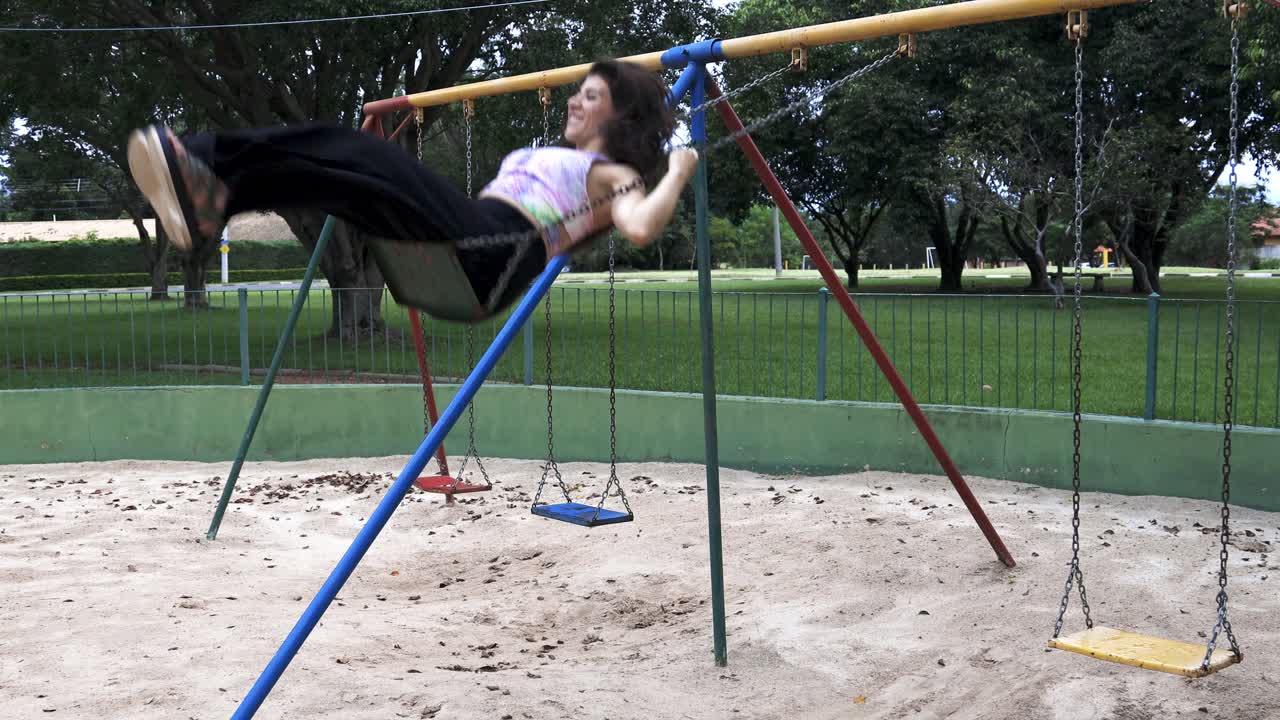 Giovane donna con gonna lunga e camicia colorata che dondola e ride, sentendosi libera e felice in un parco, ripresa lateralmente.