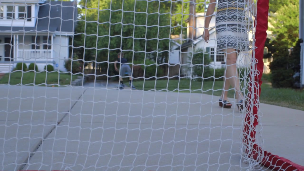 Mom walks down the driveway and out of frame as boy playing roller hockey skates.
