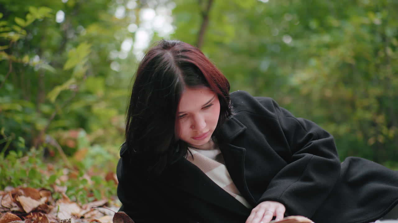 숲 바닥에 누워 책을 읽으며 차분하게 페이지를 넘기는 편안한 여성, 포근한 코트, 주변의 가을 낙엽, 조용한 야외 학습 시간, 자연 속 녹지에서 문학과 마음 챙김에 집중