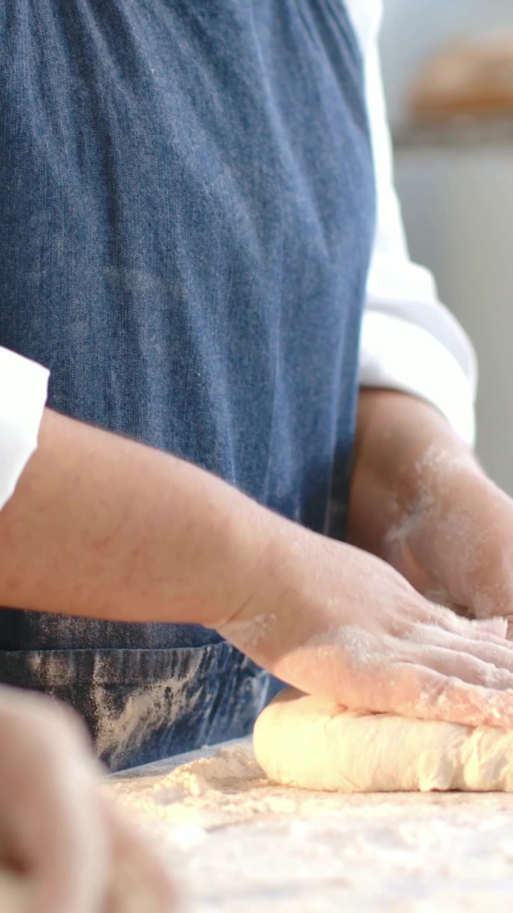 Vertical video: Shaping dough at start, denim-apron baker forming loaf on floured bench for baking