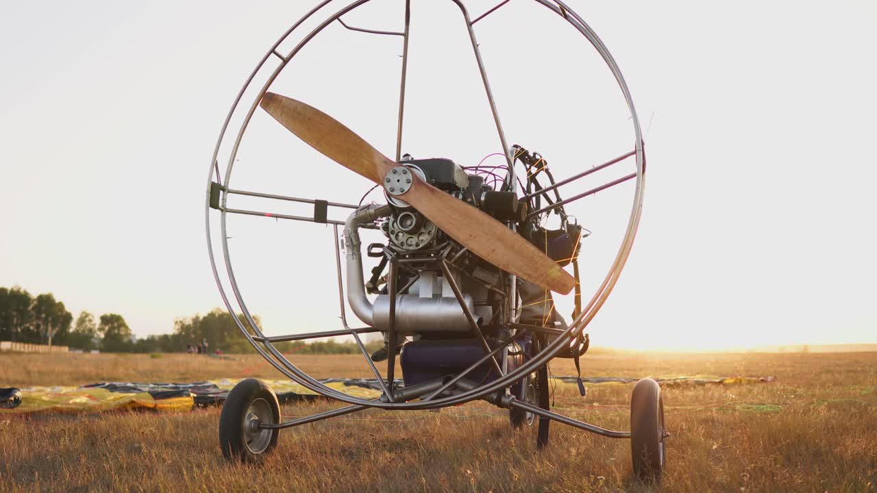 모터 패러글라이더는 해가 지는 시점에 나무 프로펠러와 함께 필드에 서 있으며, 조종사는 낙하산을 놓고 슬링을 정렬합니다. 비행 준비