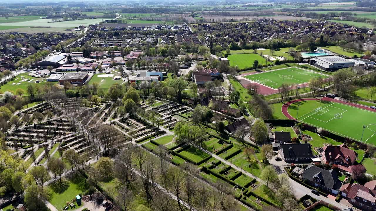 Graveyard cemetery beside green soccer field stadium in small american town at sunset time. Green landscape in spring season. Aerial lateral wide shot.