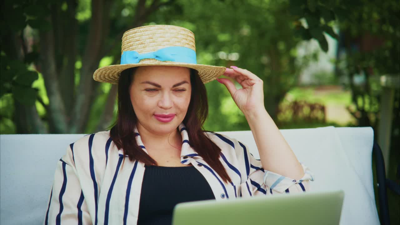 Woman wearing a straw hat, sitting outdoors with laptop, enjoying a sunny day in a garden