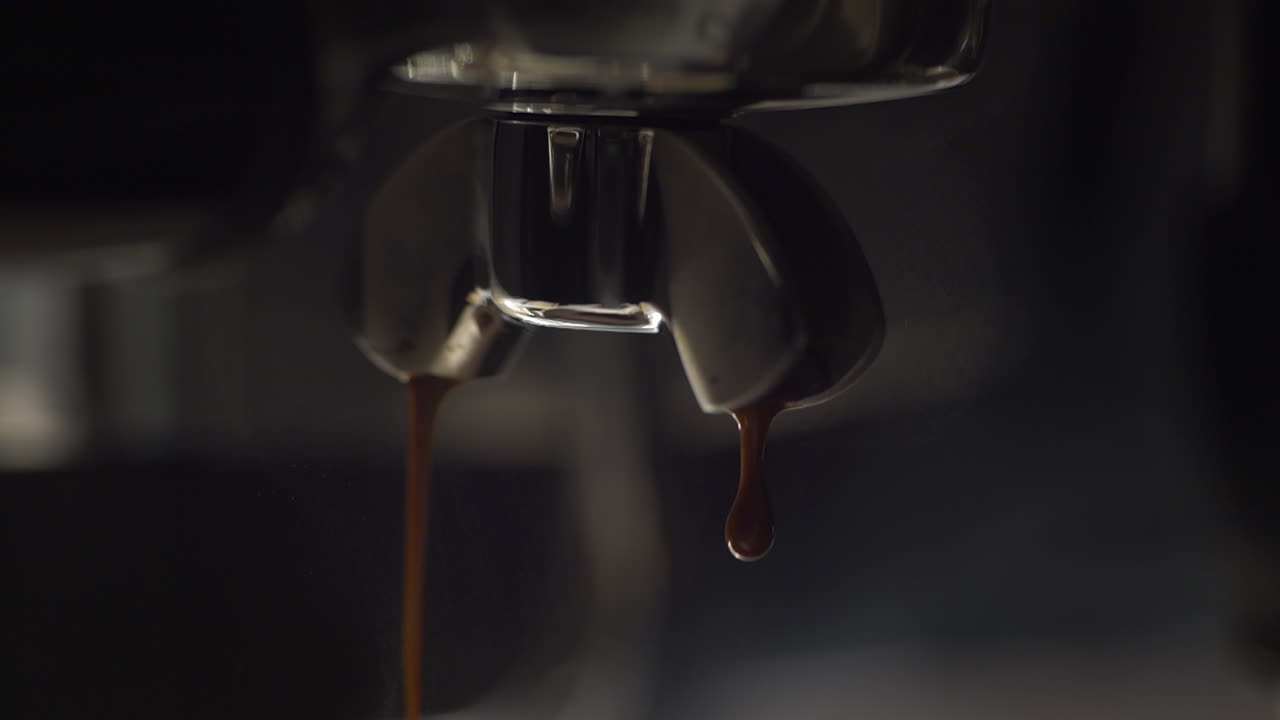Primo piano al rallentatore di caffè espresso che gocciola da un portafiltro durante l'estrazione. Crema densa si forma e scende sotto pressione, catturato con illuminazione suggestiva, forte contrasto e texture dettagliata.
