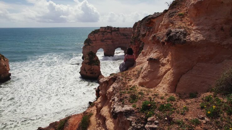 Caucasian brunette woman sitting on cliff enjoying heart shaped natural arches with sea stacks and waves crashing aerial 4k drone view of Estrada da Caramujeira beach in the Algarve region of Portugal