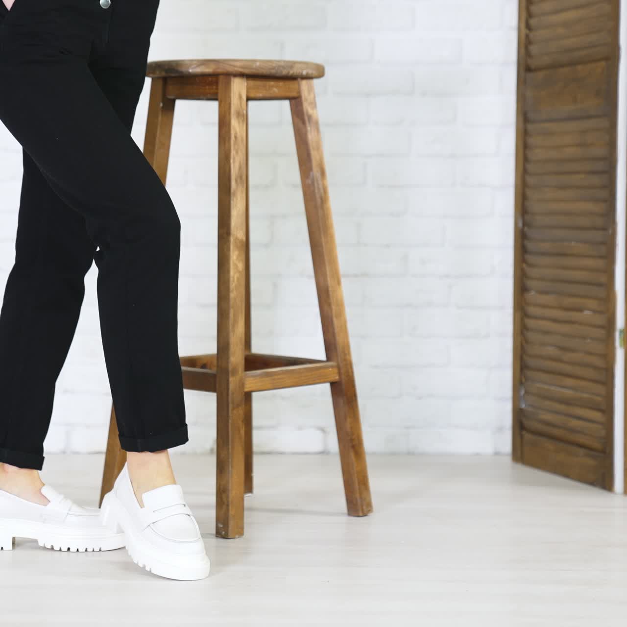 Model in black jeans demonstrating white massive shoes. Lady turns around to show the fashionable footwear. Wooden tabouret at backdrop