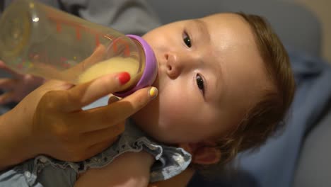 mom's hand holding the bottle with milk and feeding hungry baby holding baby in her arms, one-year-old pretty girl drinking milk from bottle - static face close-up