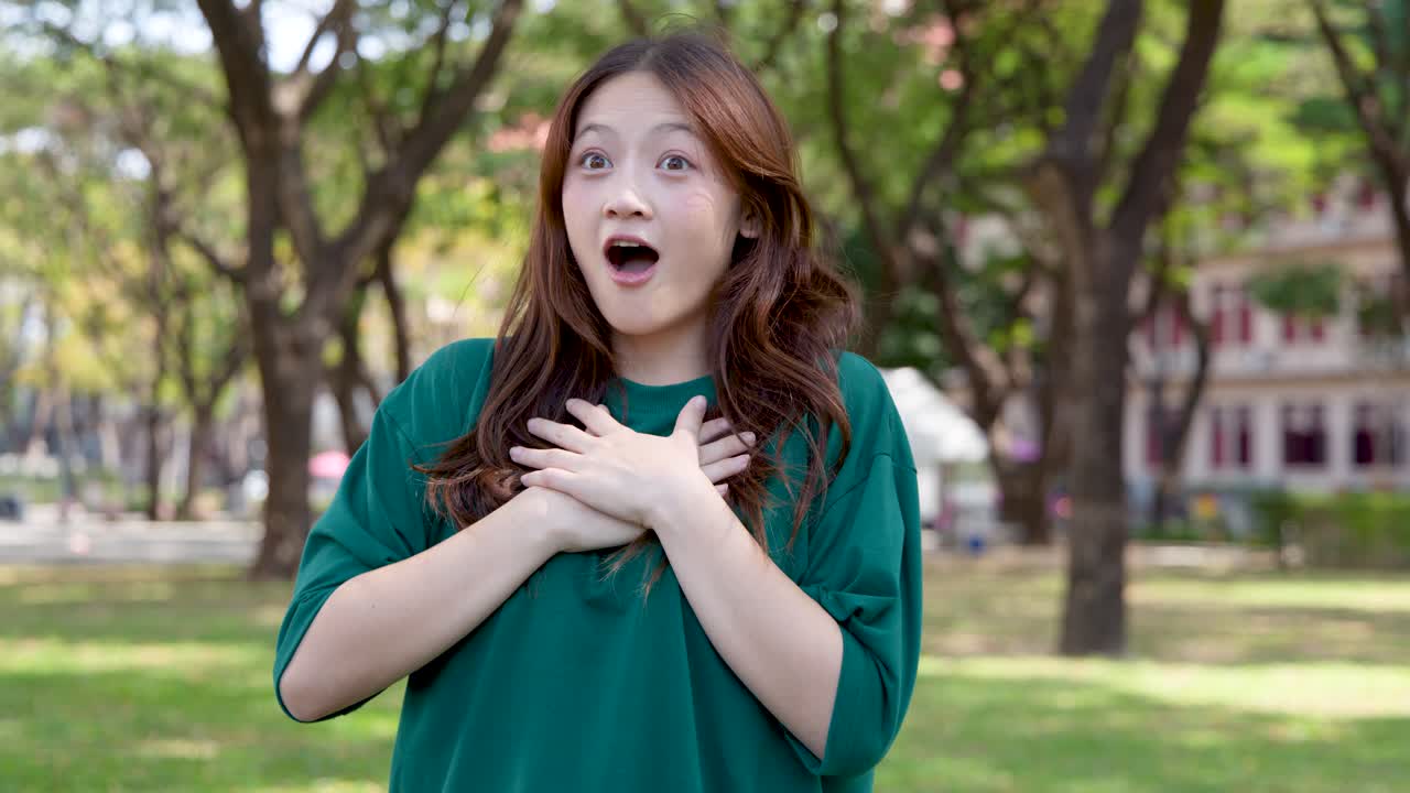 หญิงสาวชาวเอเชียที่ตื่นเต้นแสดงอาการตกใจและดีใจขณะยืนอยู่ในสวนสาธารณะที่มีแดดจ้า