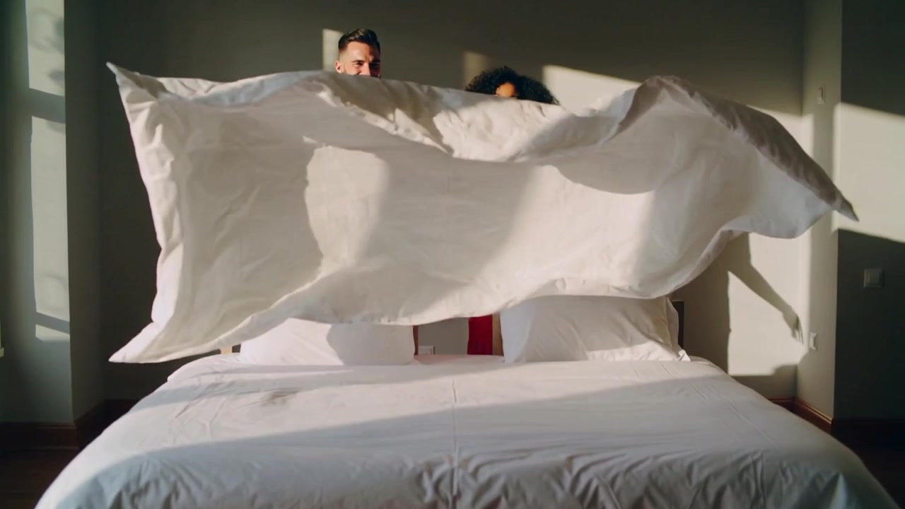 Donna in abito rosso e uomo in verde acqua che stendono il piumone, rifanno il letto in camera da letto vicino alla finestra, sistemando i cuscini.