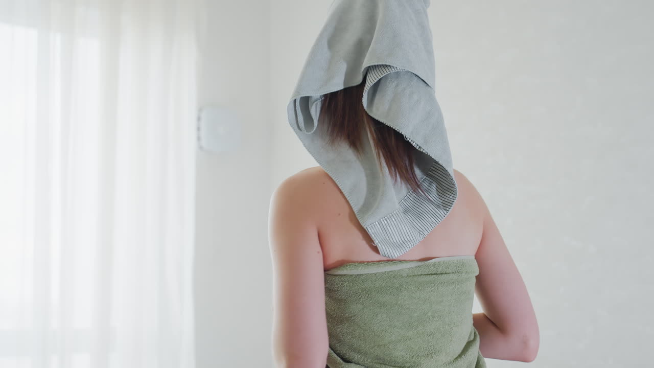Back view of woman wrapped in green towel with grey head towel standing in bright room near window and sheer curtains showing blemished skin under soft natural light and elegant chandelier overhead