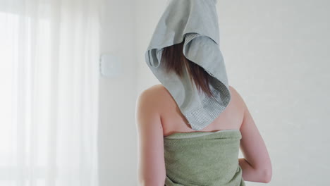 back view of woman wrapped in green towel with grey head towel standing in bright room near window and sheer curtains showing blemished skin under soft natural light and elegant chandelier overhead