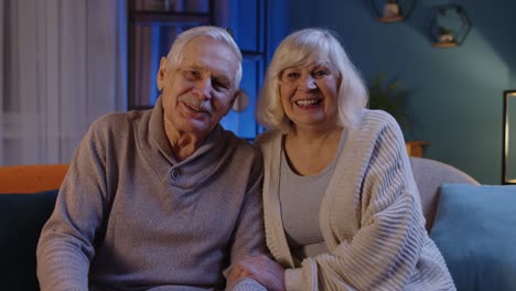 happy old senior elderly family couple hugging laughing smiling looking at camera at home sofa