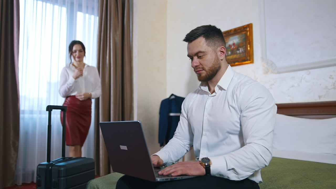 Handsome man with a laptop in hotel. Young couple arrived into a hotel. Attractive woman coming to her husband working on a laptop.