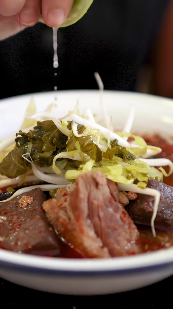 柔らかな自然光の下、北タイカレーソースがかかり、漬物とスプラウトが添えられた米麺のボウルに、手がライムを絞りかけている。