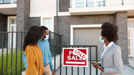 Joven-Pareja-Afroamericana-Feliz-Con-Máscaras-Médicas-Comprando-Casa-En-Las-Afueras-Y-Hablando-Con-Una-Agente-Inmobiliaria-Que-Sostiene-Un-Dispositivo-De-Tableta-En-Las-Manos