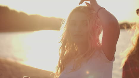 una chica rubia con camiseta blanca, maquillaje brillante y largos pendientes está moviendo su cuerpo y manos en la fiesta de playa en la costa del río de arena con cerveza.