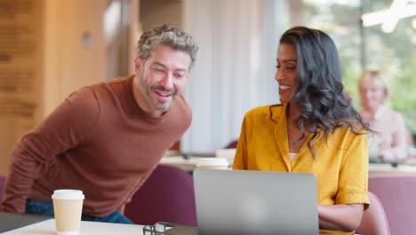 Mature-Male-Businessman-Brings-Takeaway-Coffee-For-Female-Colleague-As-They-Have-Meeting-With-Laptop