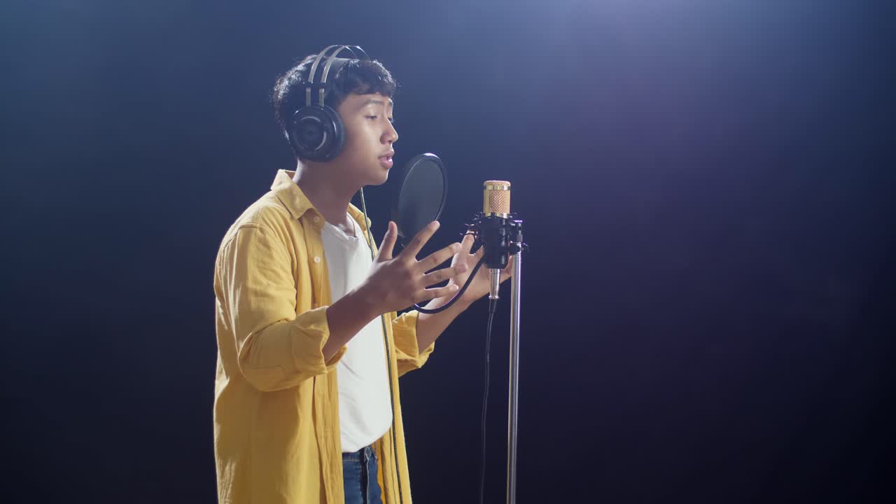 흑색 배경 에 있는 콘덴서 마이크 로 노래 하는 헤드폰 을 착용 한 젊은 아시아 소년 의 측면 시각