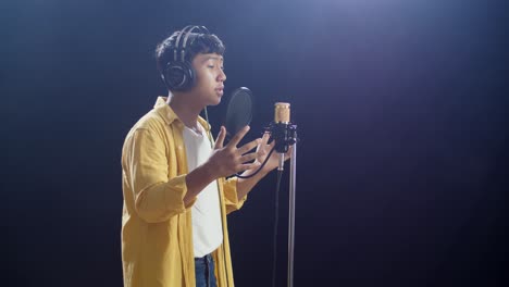 vista lateral de un joven asiático con auriculares cantando en un micrófono condensador en el fondo negro