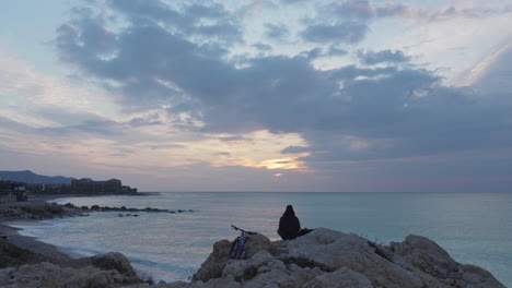 Der-Zeitraffer-Fängt-Den-Sonnenaufgang-über-Einem-Einsamen-Mann-Ein,-Der-Auf-Felsen-Meditiert-Und-Ins-Meer-Und-In-Die-Wolken-Blickt