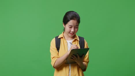 asian woman student with a backpack smiling and taking note on the tablet while standing in the green screen background studio