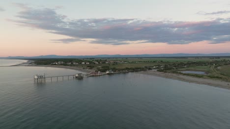 Vista-Aérea-En-Cámara-Lenta-De-Cabañas-De-Pesca-A-Orillas-Del-Estuario-Al-Atardecer,-Máquina-De-Pesca-Italiana,-Llamada-&quot;trabucco&quot;-Ravenna-Cerca-Del-Valle-De-Comacchio