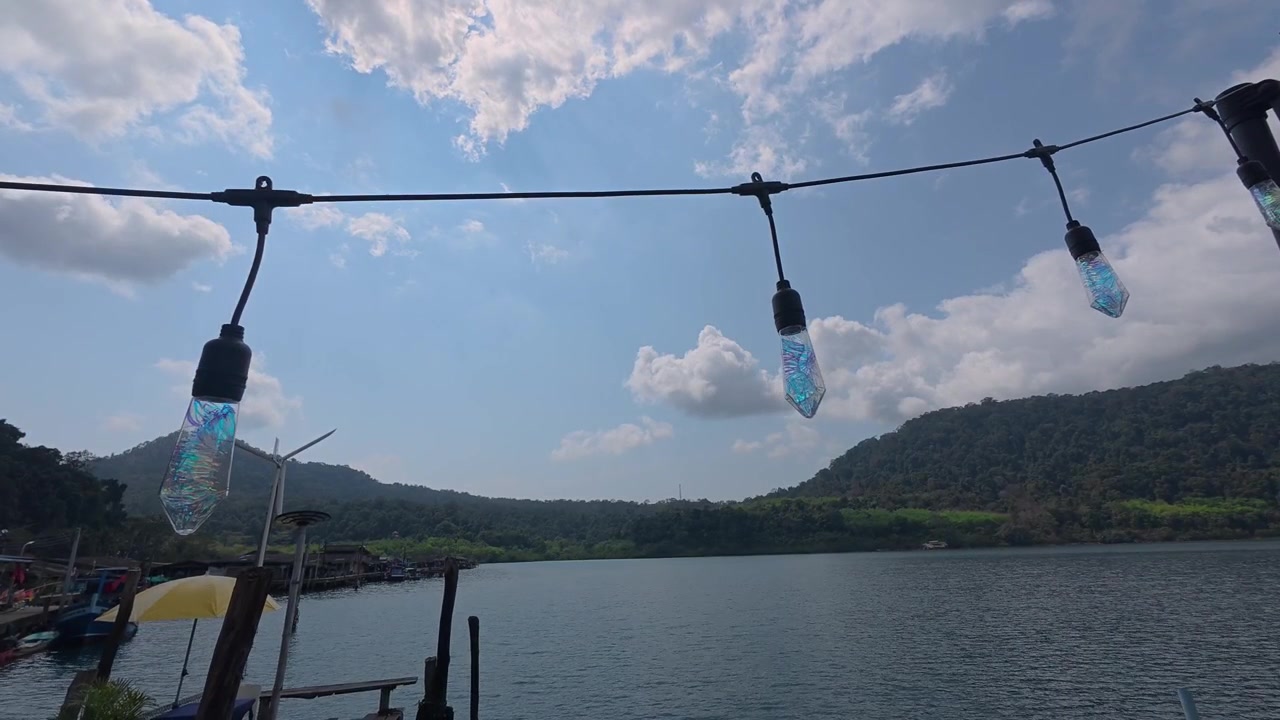 Riprese orizzontali che si muovono a sinistra e poi panoramicano a sinistra lungo il lungomare del villaggio di pescatori di Ao Yai, con luci decorative, viste di una baia calma e colline boscose sull'isola di Koh Kood, Thailandia