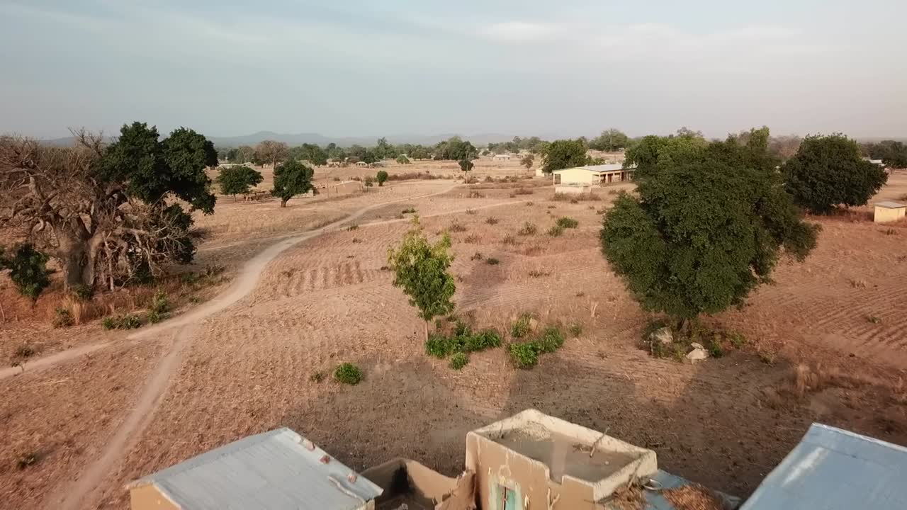 Primo piano aereo di un drone su un tradizionale complesso familiare del Ghana settentrionale con pareti di fango, paglia e tetti di zinco, circondato da terreni agricoli aridi e alberi di baobab
