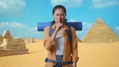 asian female hiker with mountaineering backpack looking at camera and making shh gesture while traveling in pyramid of giza