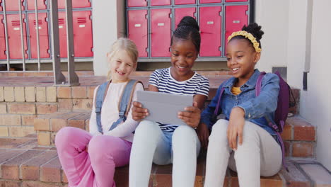 in school, three girls sitting on steps using tablet, smiling and laughing