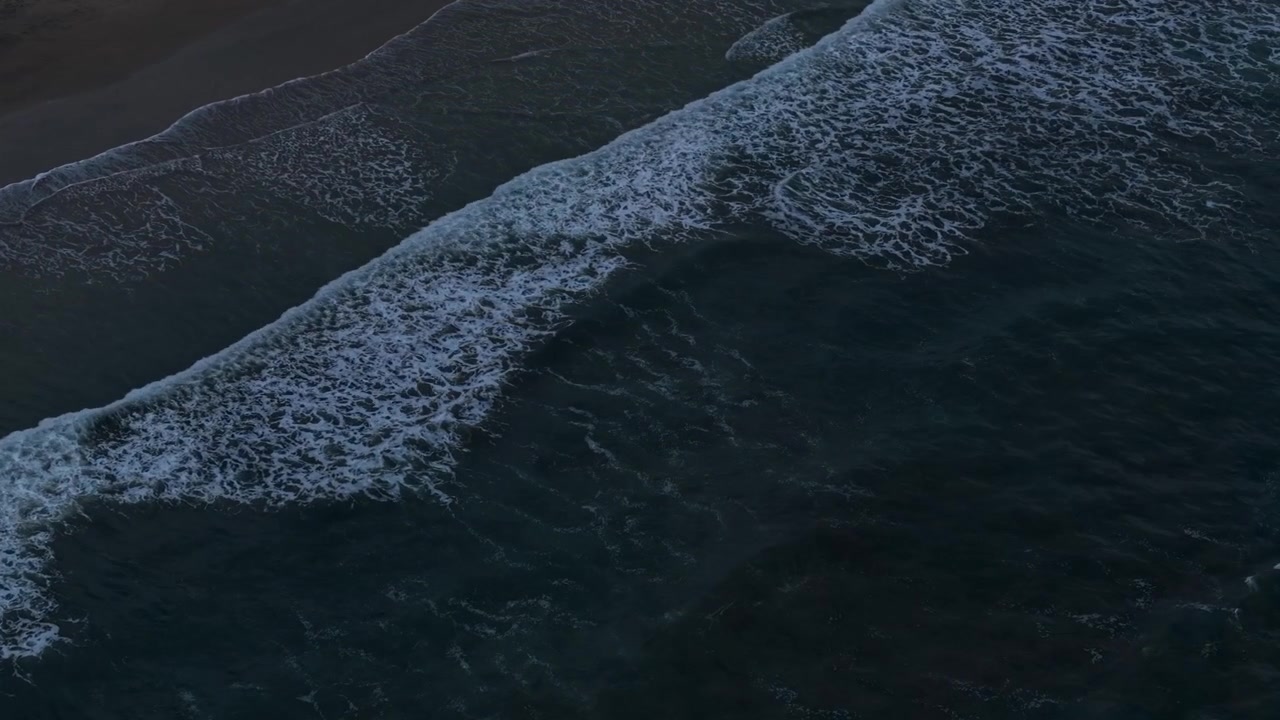 Top down aerial view of dark ocean waves with white foam gently breaking on a sandy shore at night