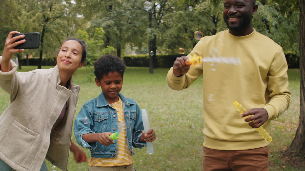 feliz familia afroamericana soplando burbujas de jabón y tomando selfies en el parque