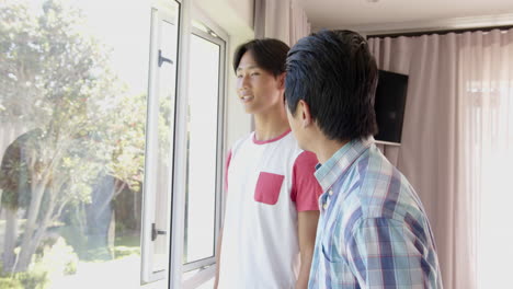 asian father and teenage son smiling and talking near window in a bright living room