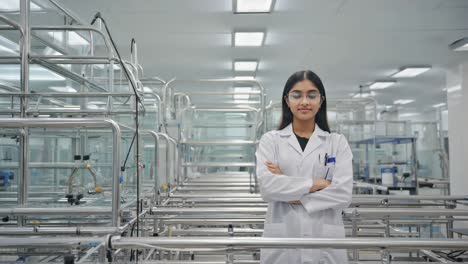 young scientist wearing lab coat and protective glasses standing with arms crossed in pharmaceutical factory, advanced equipment in background ensuring quality and innovation in medicine production
