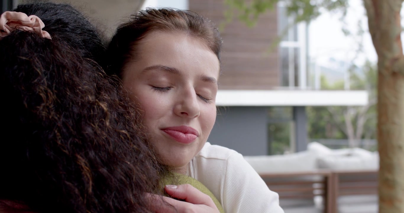 Amiche sul balcone, donna con top bianco che si porta una mano al petto, parlando, abbracciandosi per consolare